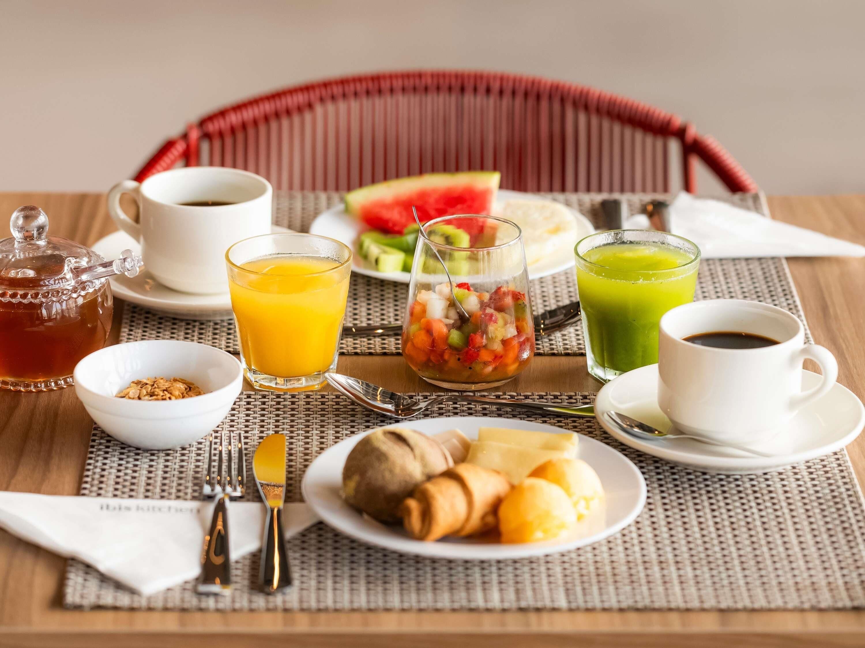 Un desayuno con una silla de mimbre, una mesa con un mantel individual, un vaso de jugo de naranja, un vaso de jugo verde, una taza de café, un plato con croissants, un bol de granola, un vaso de agua y una rebanada de sandía.