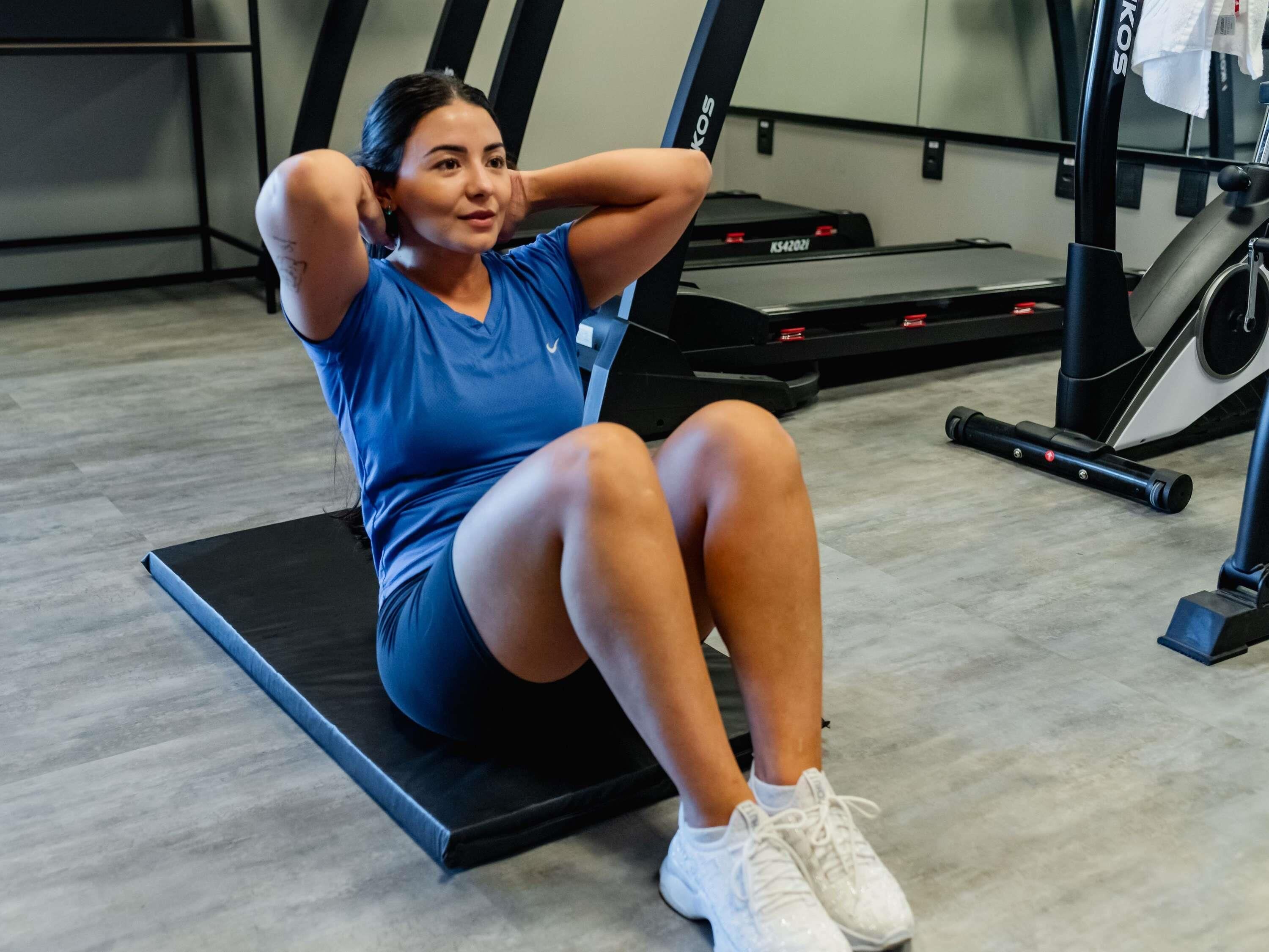 Una persona haciendo ejercicio sobre una colchoneta negra en un gimnasio, con equipo de entrenamiento al fondo.