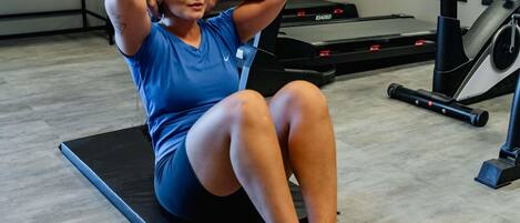 Una persona haciendo ejercicio sobre una colchoneta negra en un gimnasio, con equipo de entrenamiento al fondo.