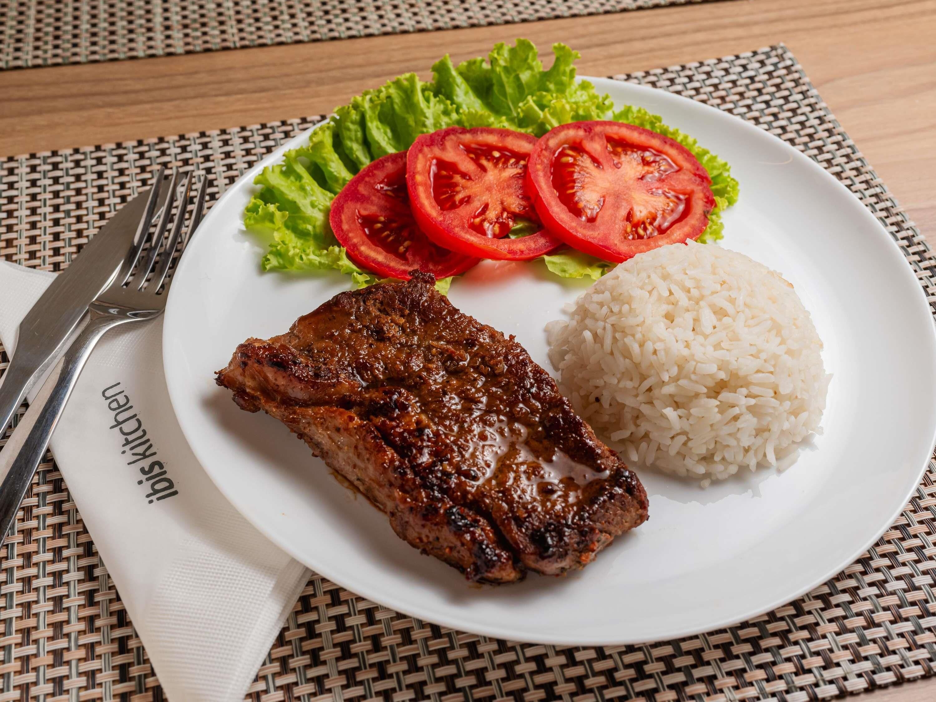 A plate with grilled meat, rice, and fresh tomato slices on a woven placemat.