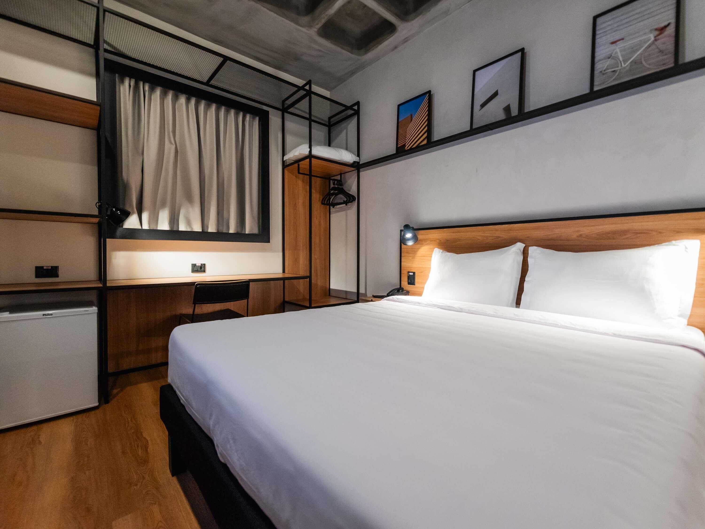 Habitación de hotel con cama, escritorio, silla, refrigerador pequeño y estante con fotografías.