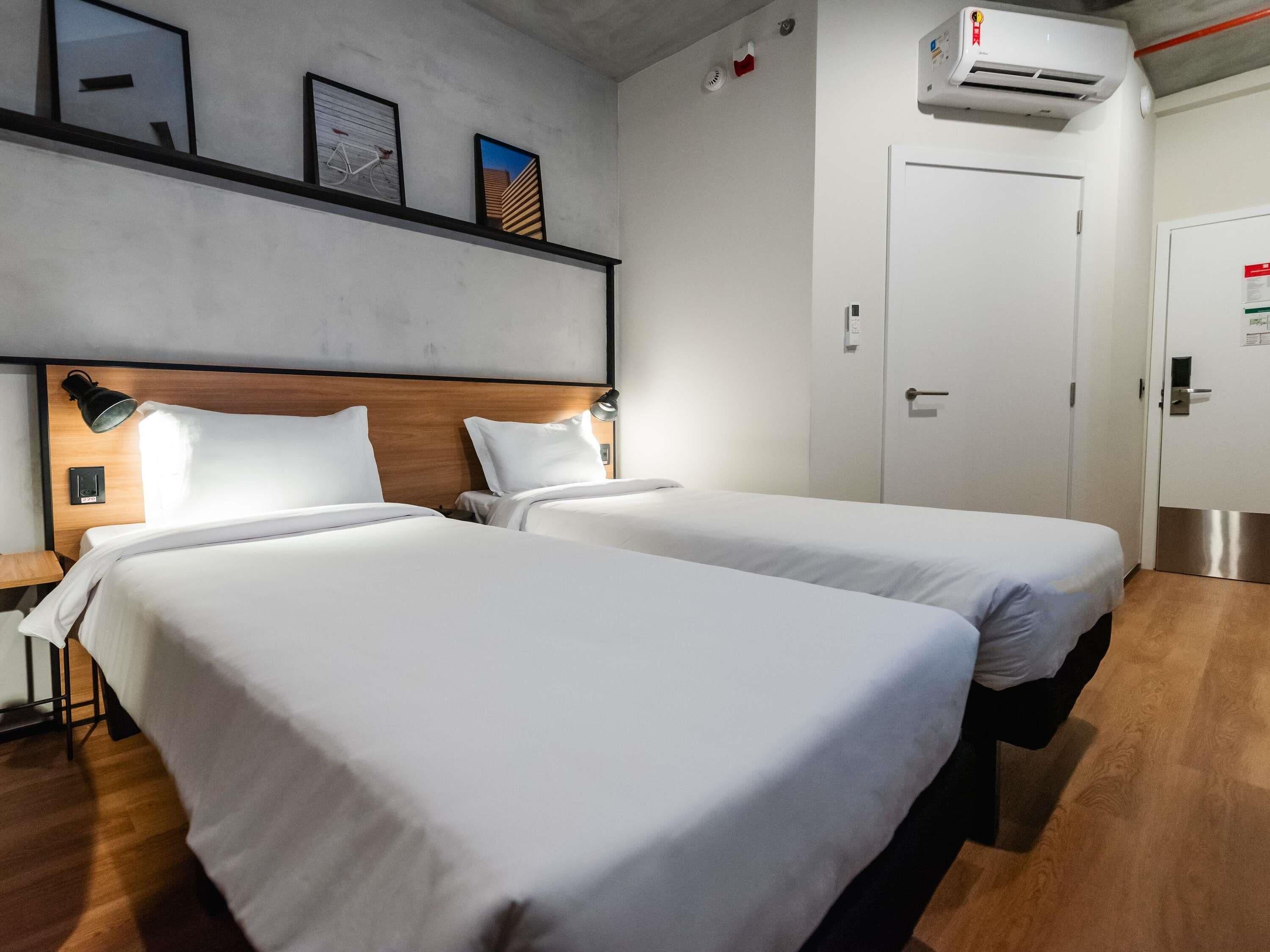 A hotel room with two beds, a wooden headboard, a mounted air conditioner, and a door with a handle.
