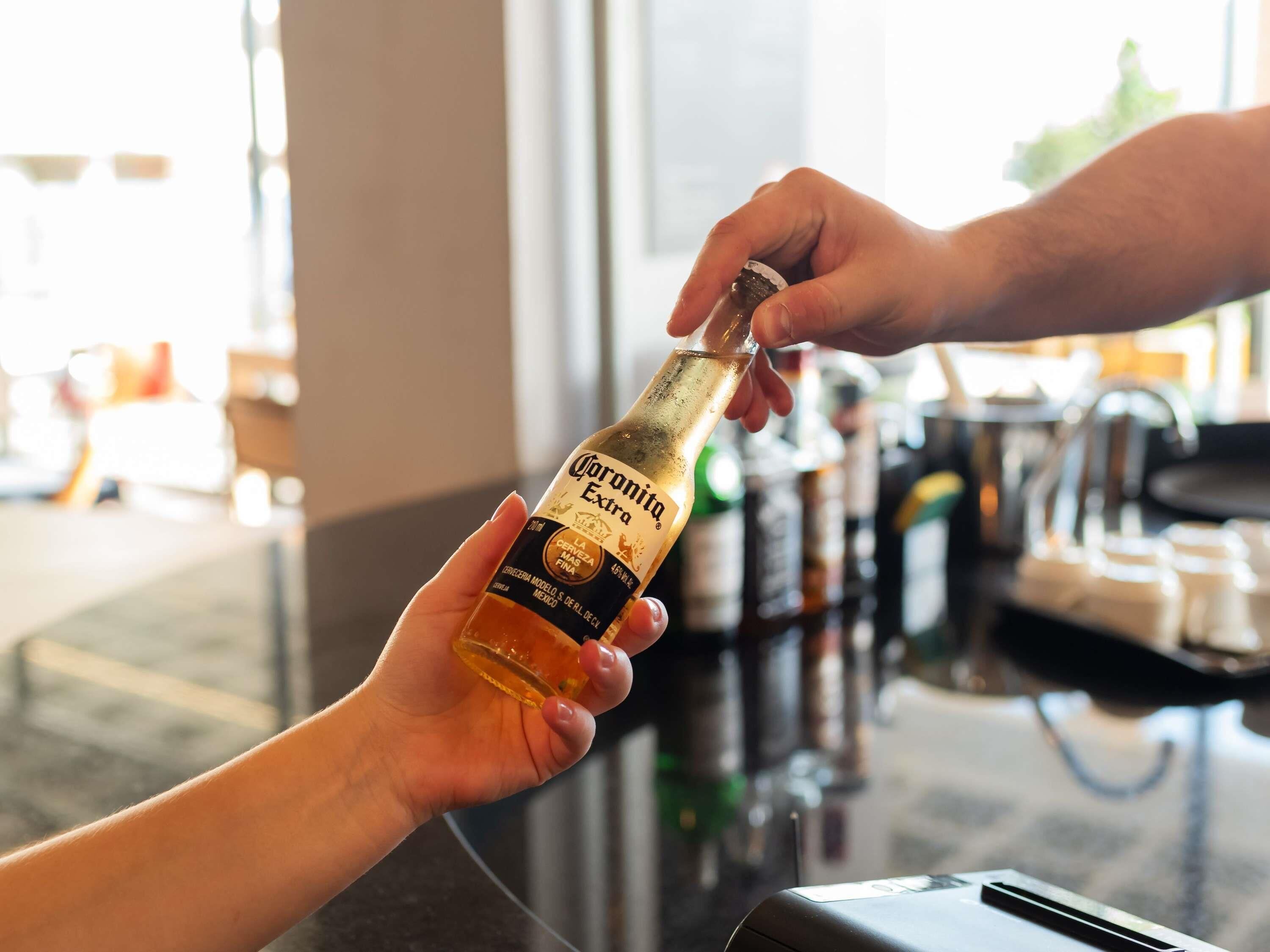 A hand holding a Corona Extra beer bottle, with another hand reaching to take it.