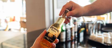 Una mano sostiene una botella de cerveza Corona Extra, mientras otra mano se extiende para tomarla.