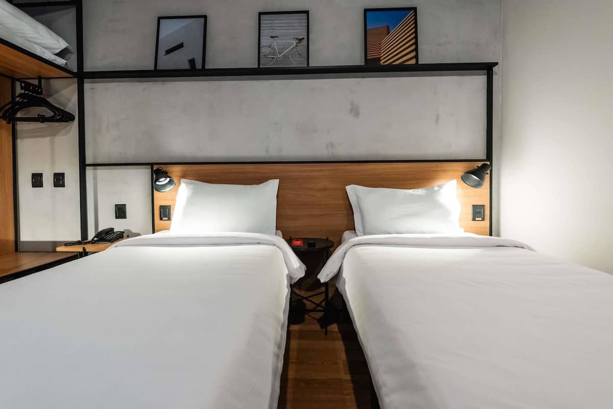 A hotel room with two beds, a wooden headboard, bedside lamps, a phone, and a shelf with a bicycle.