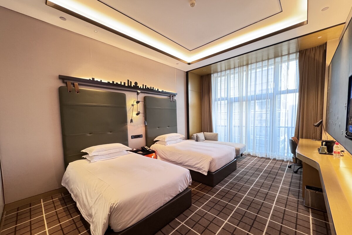 A hotel room with two beds, a desk, a chair, a TV, and a large window with curtains.