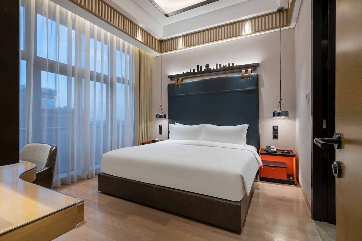 A modern hotel room with a large bed, a desk, and a chair. There is a nightstand with a lamp and a clock. The room has a city skyline view through the window.