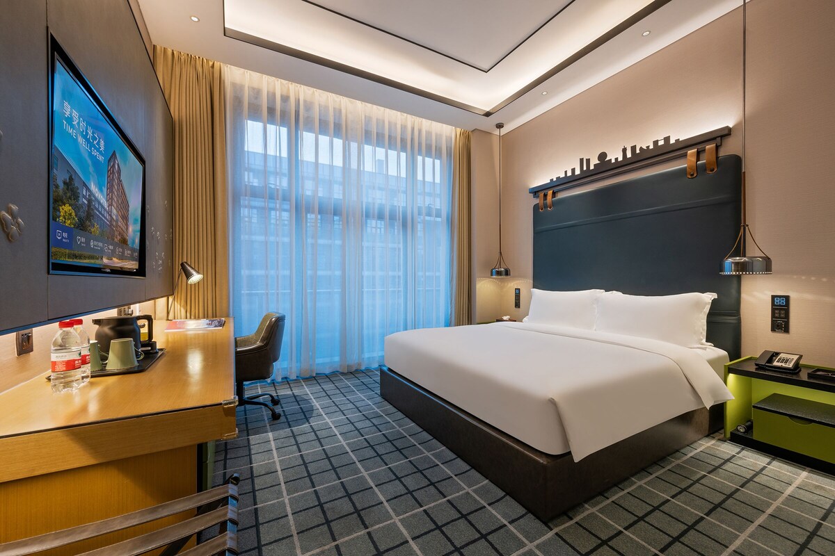 A hotel room with a large bed, a desk with a computer, a TV, and a window with curtains.