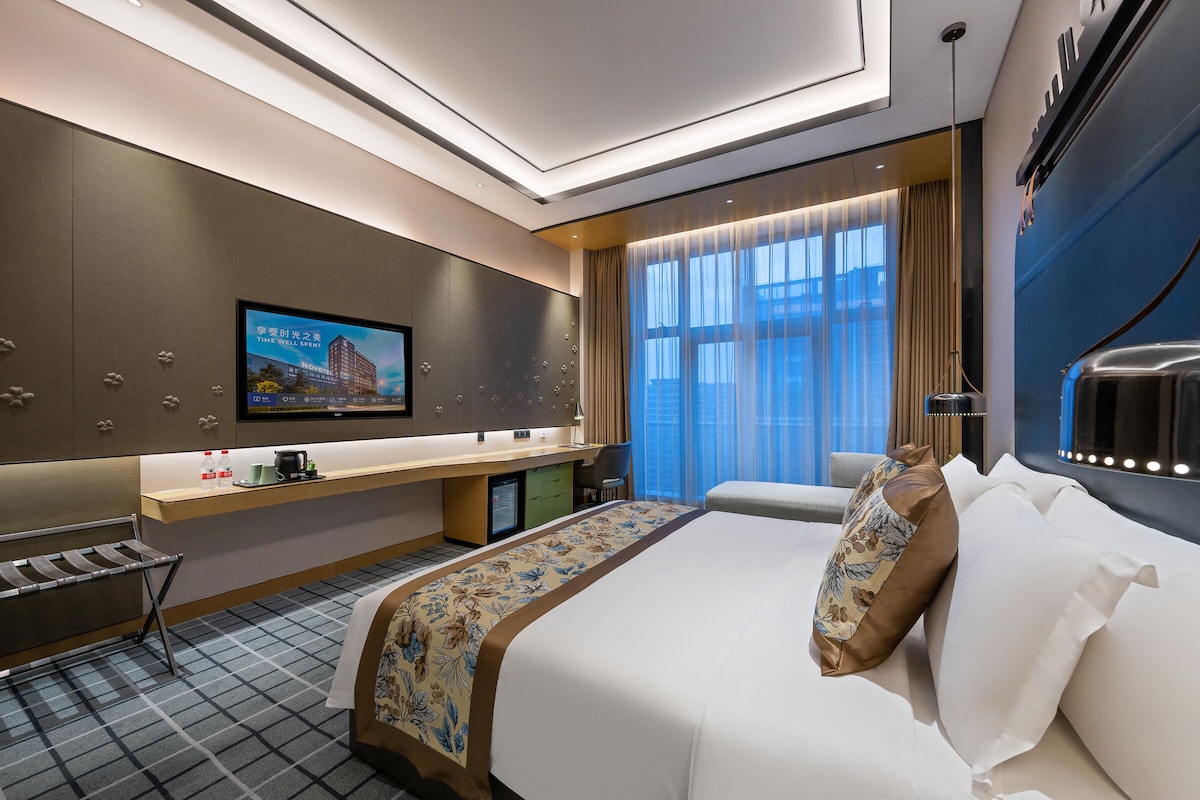 A modern hotel room with a large bed, a flat-screen TV mounted on the wall, a desk with a computer, and a window with curtains.