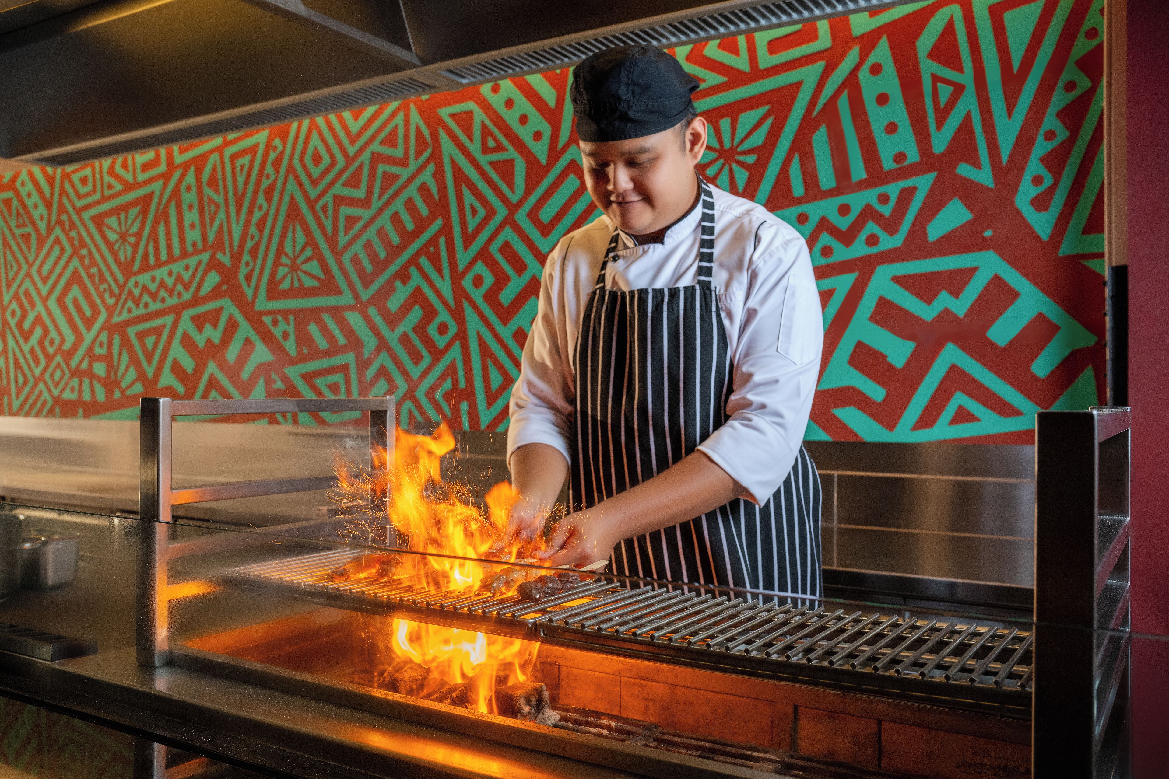 A chef grilling food on a commercial grill in a kitchen with a patterned wall.