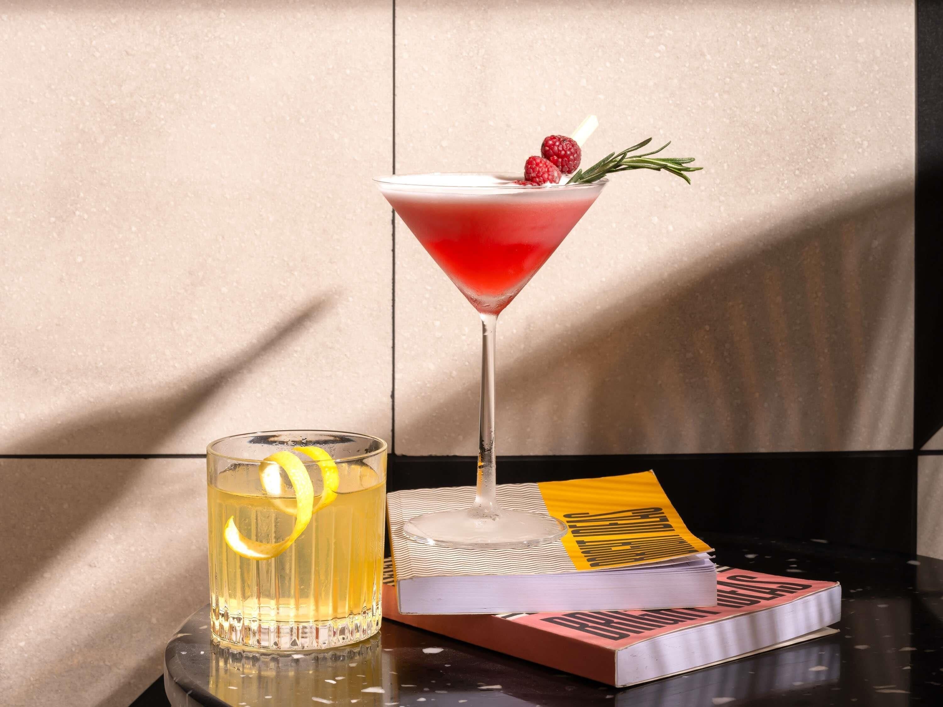 A martini glass with a red drink, a glass of yellow beverage with a lemon slice, and a book titled 'The Art of War' on a reflective surface.