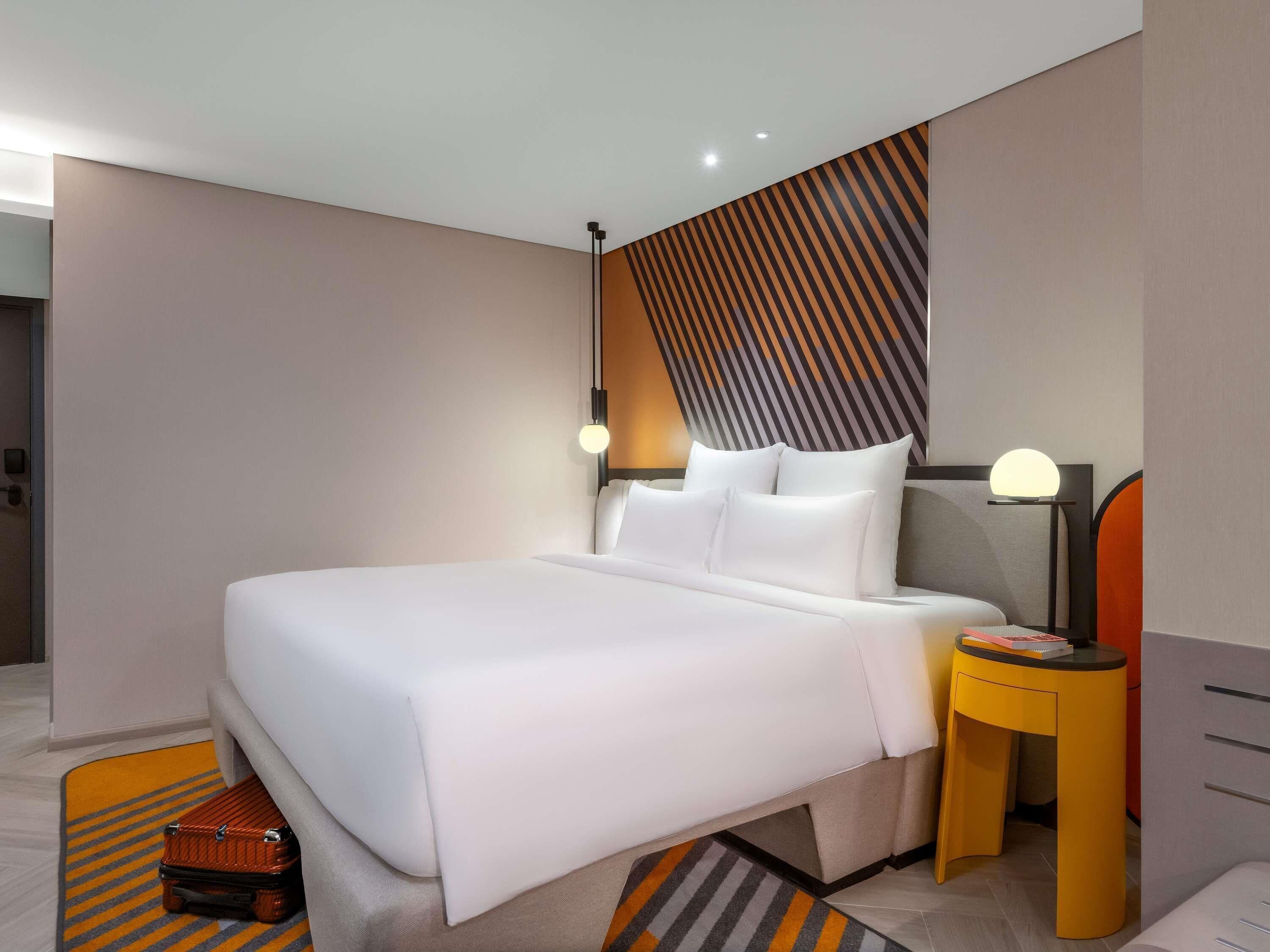 A modern hotel room with a large bed, a bedside table with a lamp, and a suitcase on the floor.