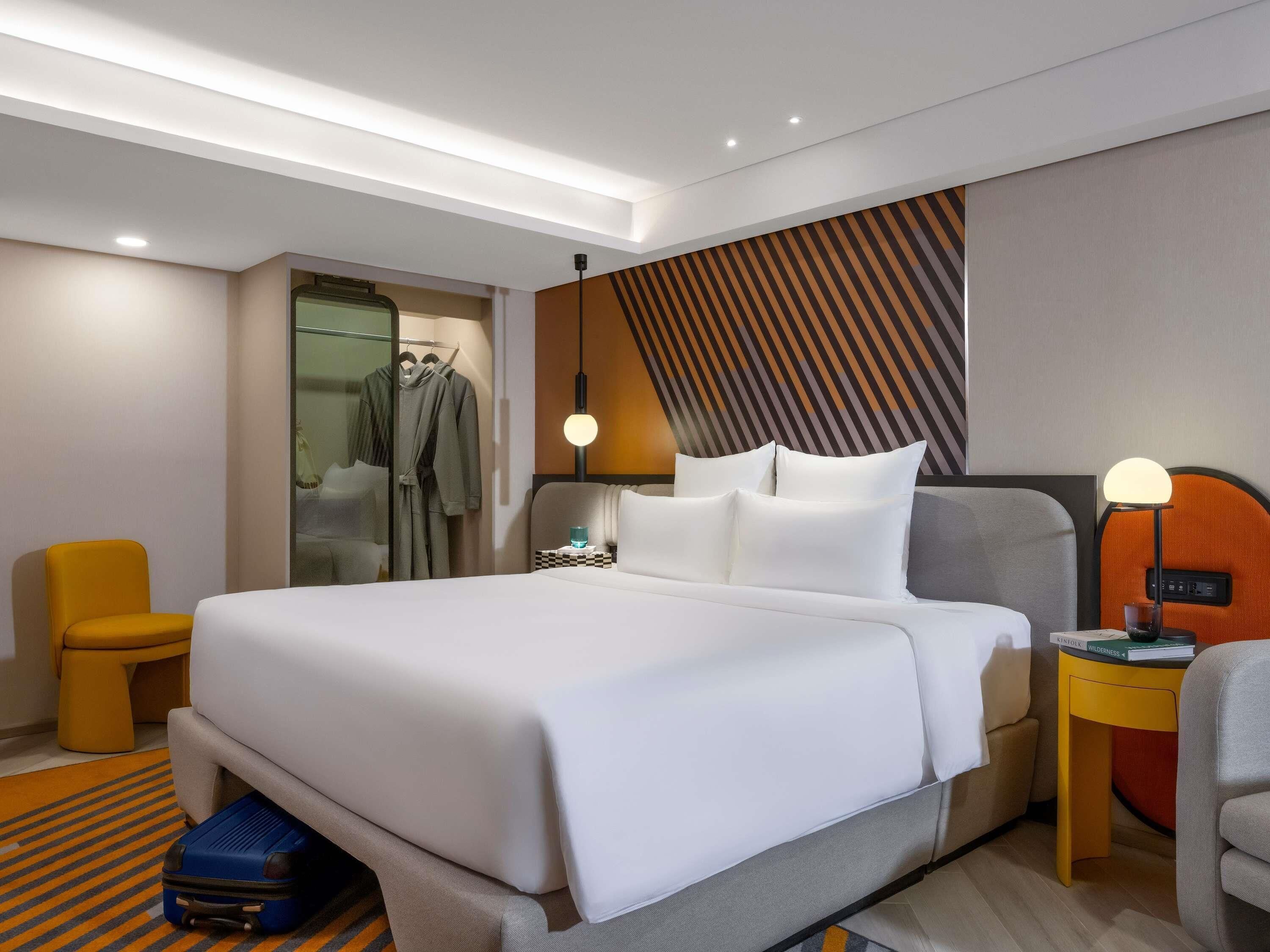 A modern hotel room with a large bed, a seating area with a chair, a small table with a lamp, and a wardrobe.
