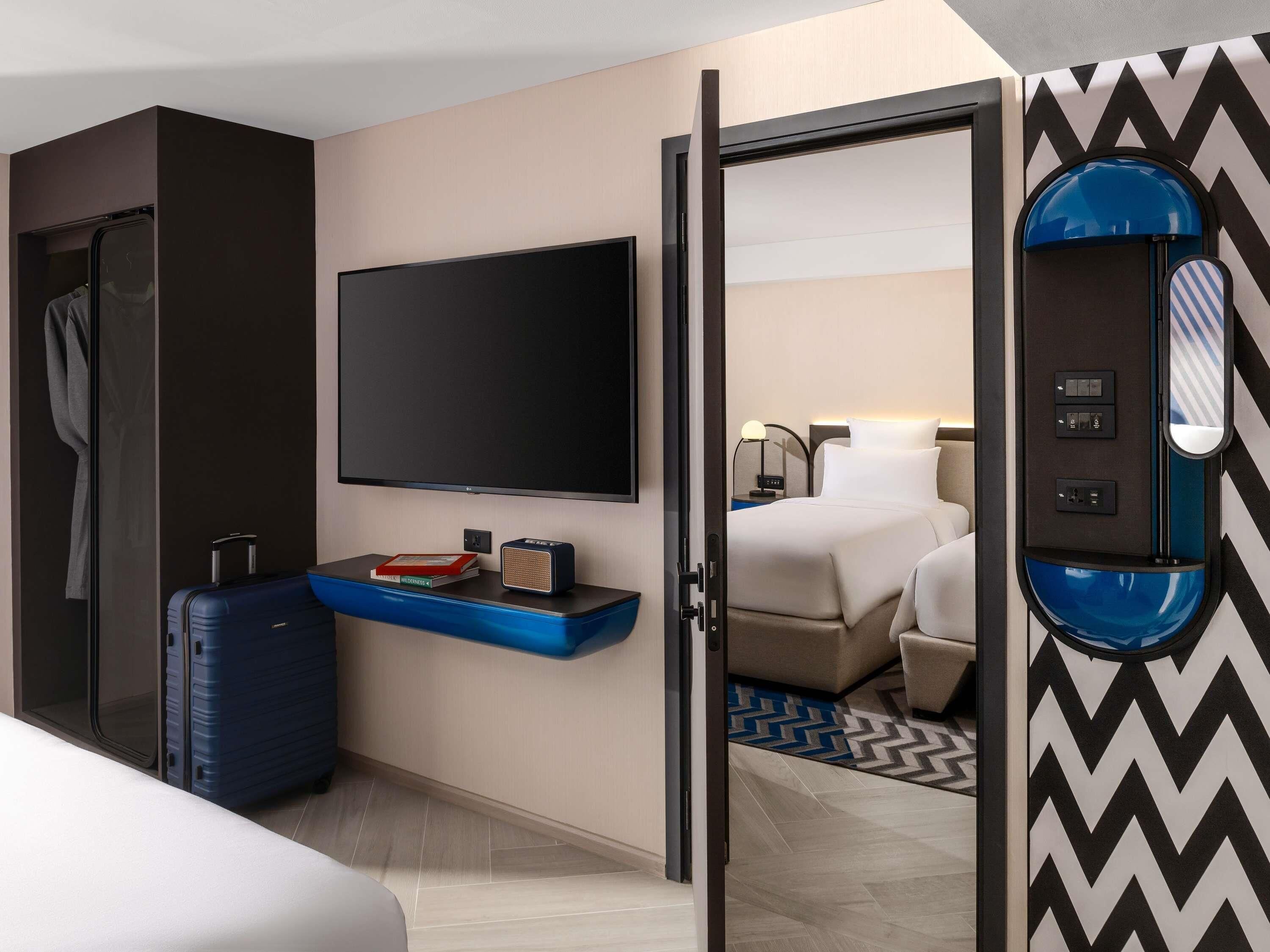 A hotel room with a large flat-screen TV, a desk with a laptop, a suitcase, and a bed with a blue and white patterned rug.