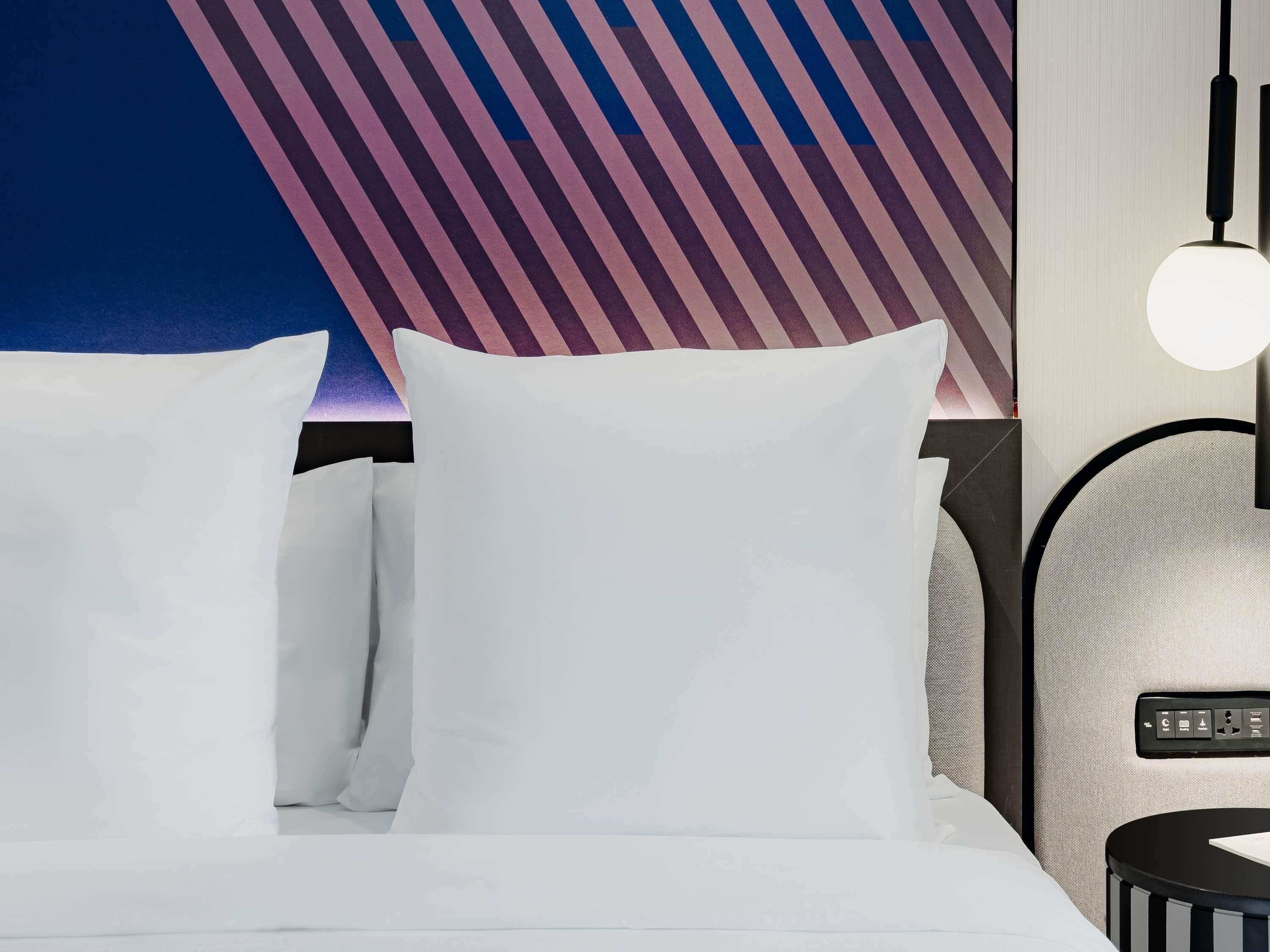 A hotel room with a bed, pillows, a nightstand, a lamp, and a wall with a striped pattern.
