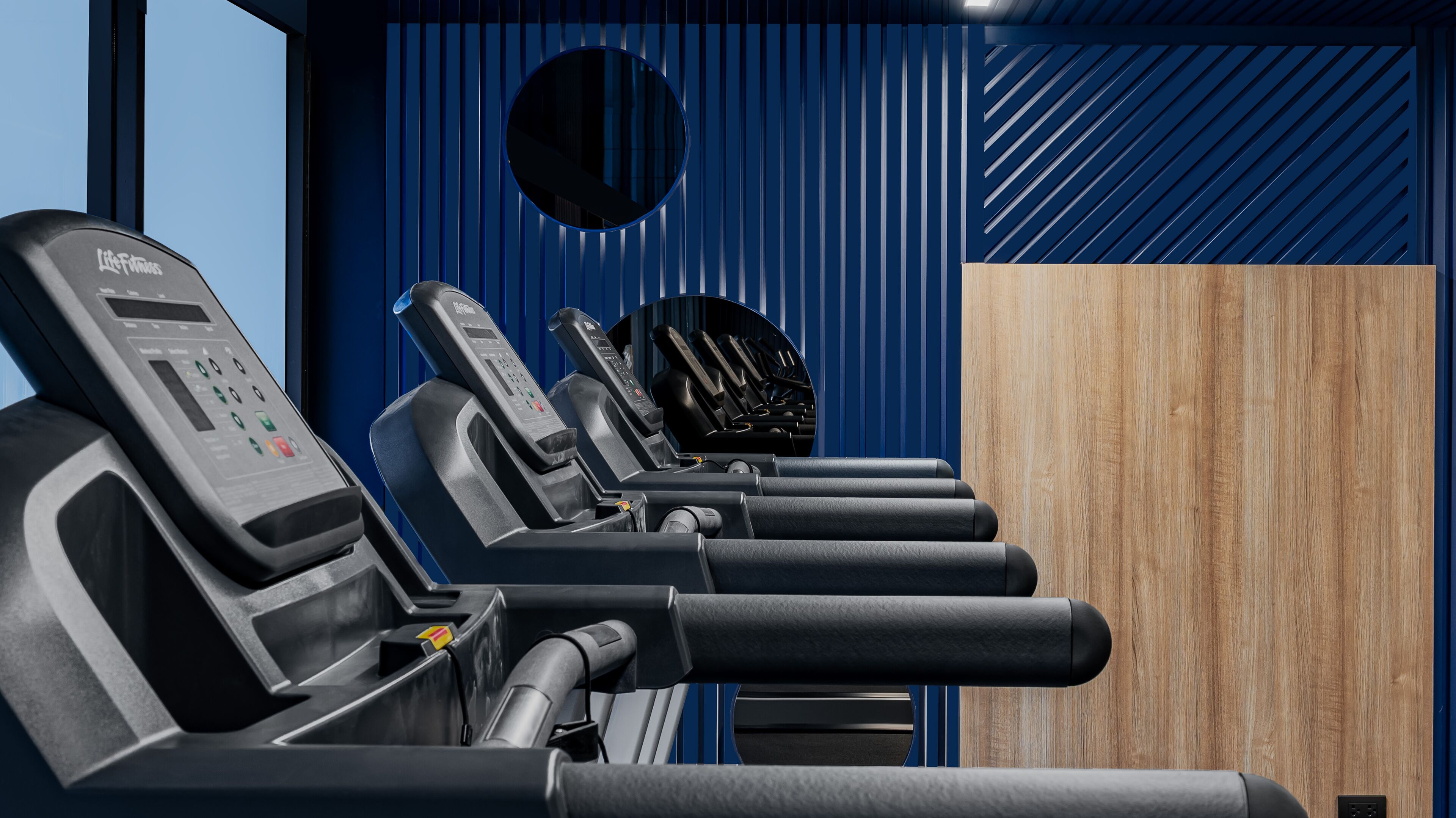 A row of exercise machines with control panels, a circular mirror, and a wooden panel in a modern room.