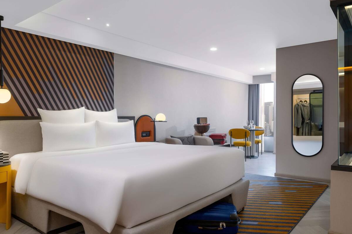 Habitación de hotel moderna con una cama grande, un escritorio, una silla, una mesita, un espejo y vistas a la ciudad.