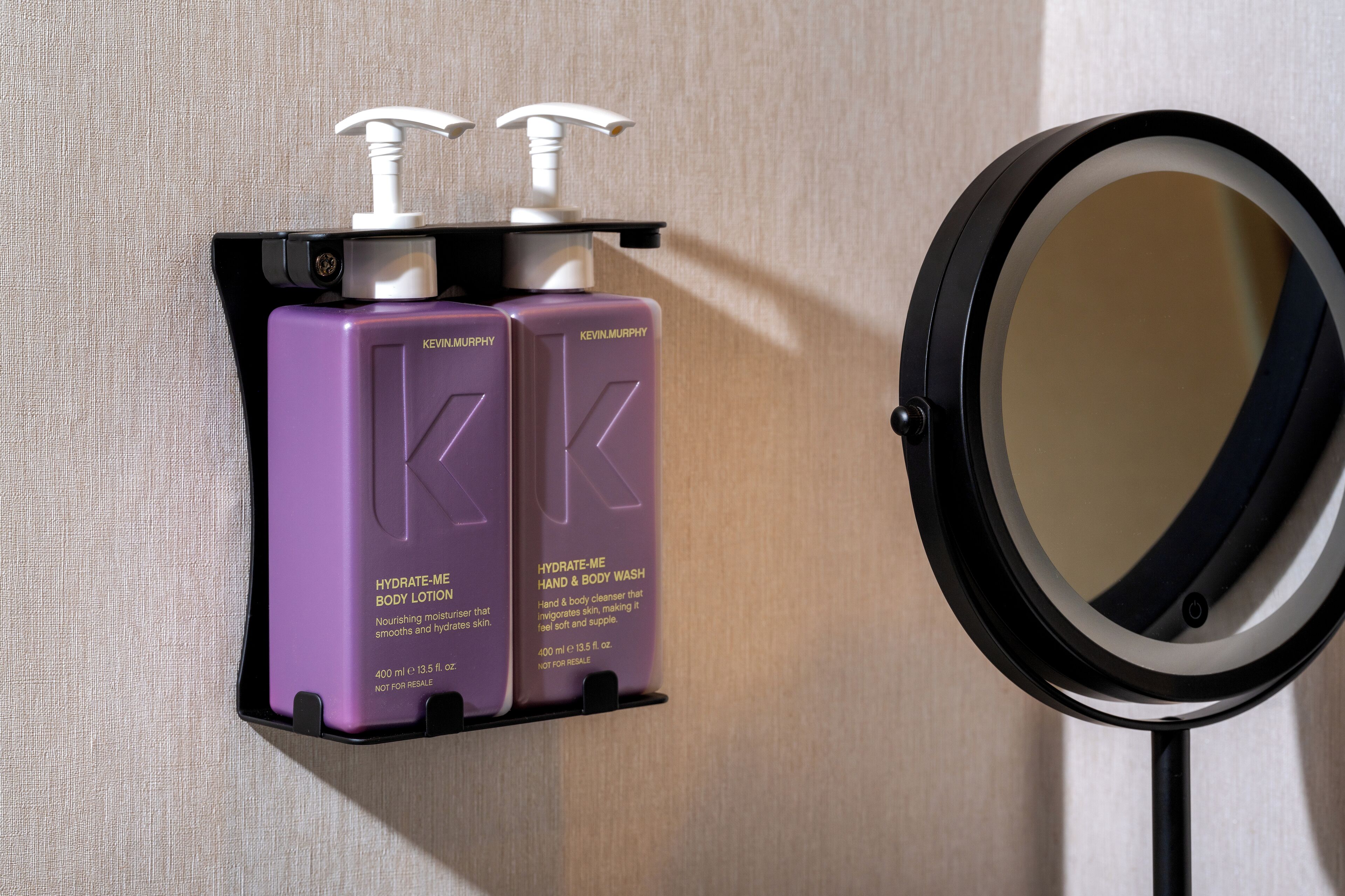 A bathroom with two purple Kevin Murphy hand and body wash dispensers mounted on a black holder, and a round mirror with a black frame.