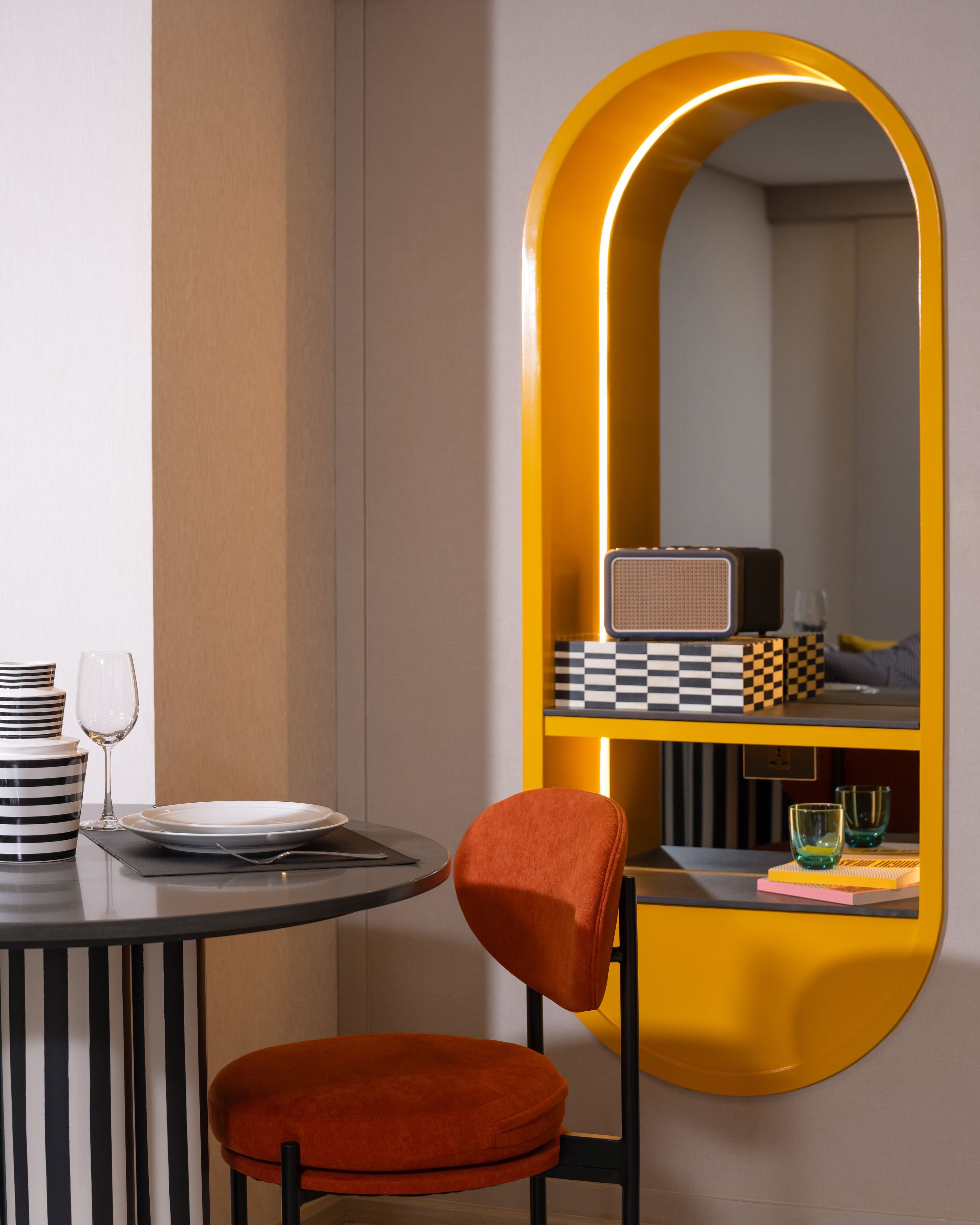 Una habitación moderna con una mesa redonda, una única silla naranja, un espejo con marco amarillo y un patrón a cuadros en la pared.