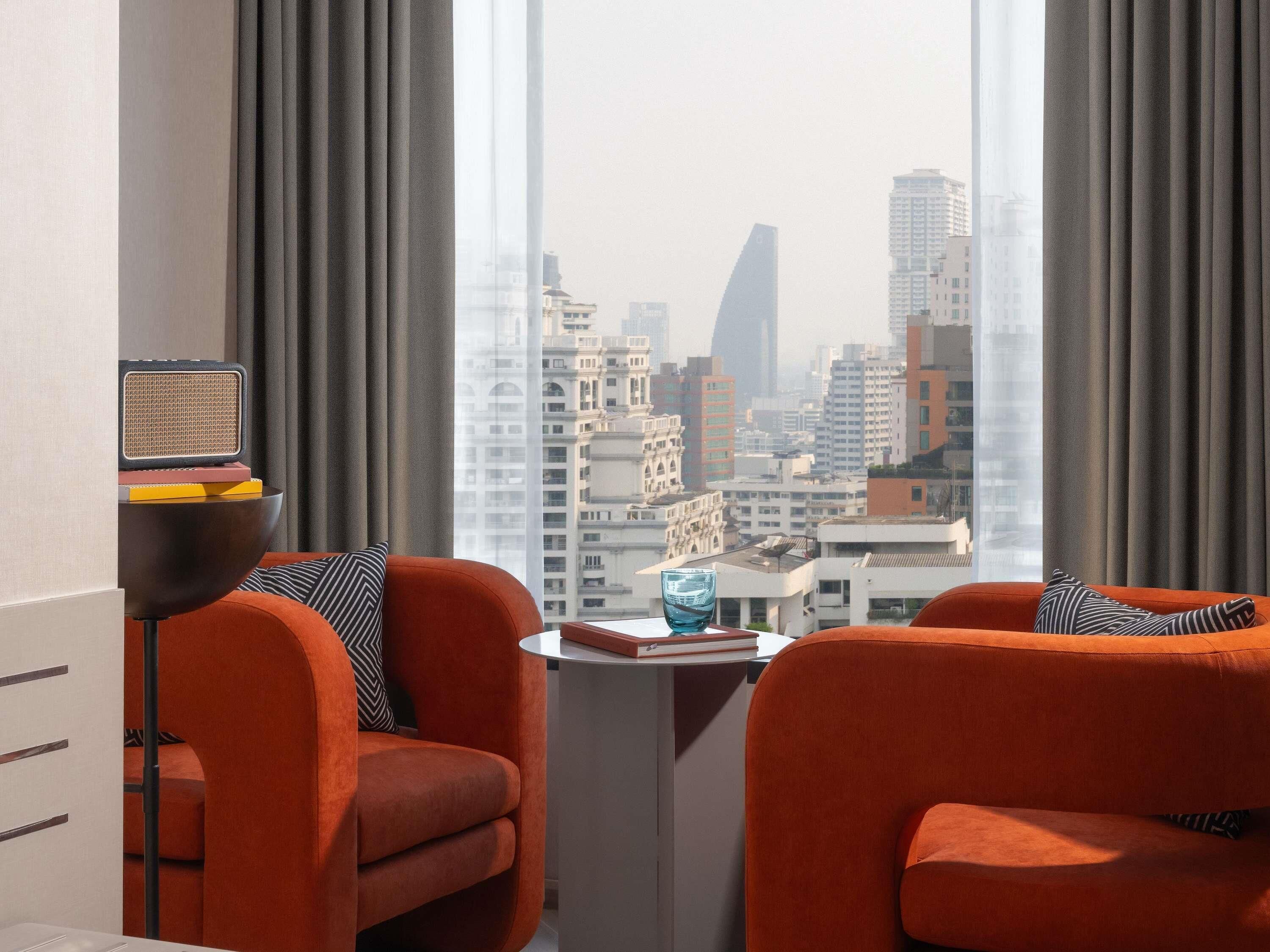 Un salón moderno con sofás naranja, una mesita y vistas a un paisaje urbano a través de amplios ventanales.