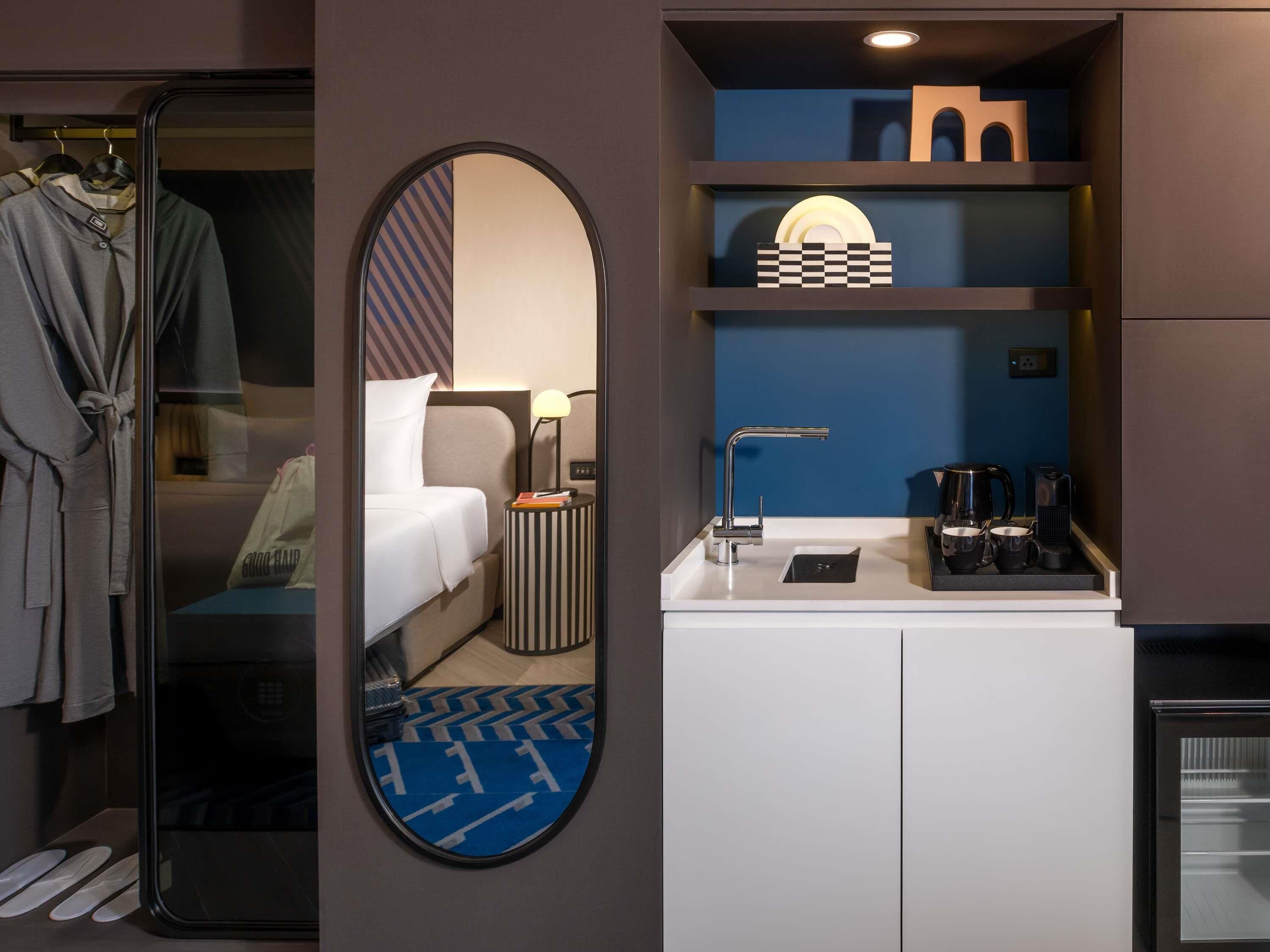 Habitación de hotel con cama, espejo, armario, mesita de noche con lámpara, cafetera y pequeña estantería con objetos decorativos.
