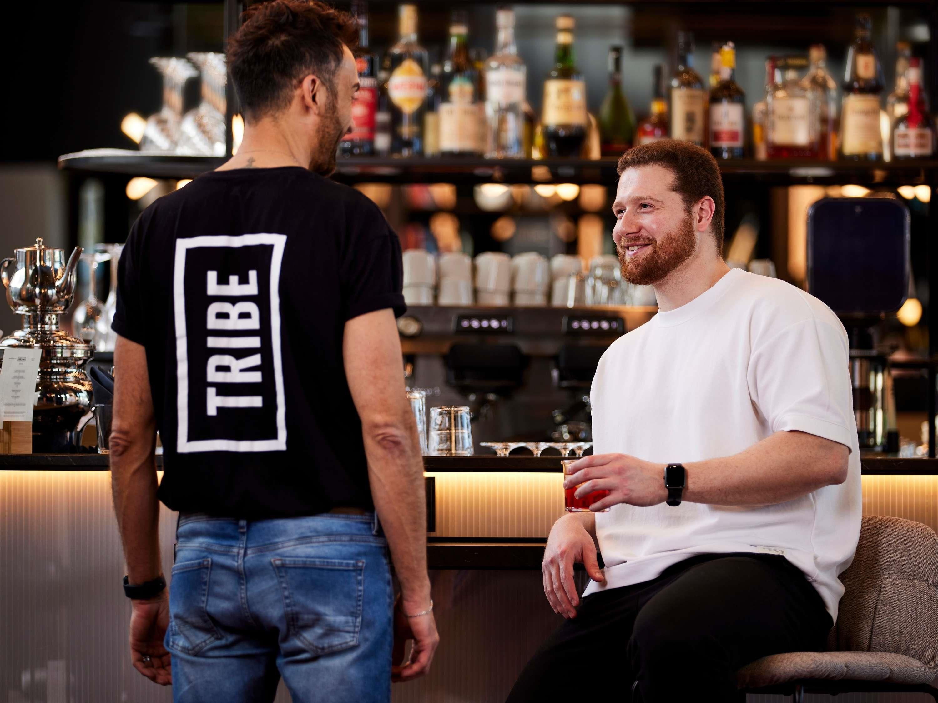Two men in a bar setting with a variety of bottles and a coffee machine in the background.