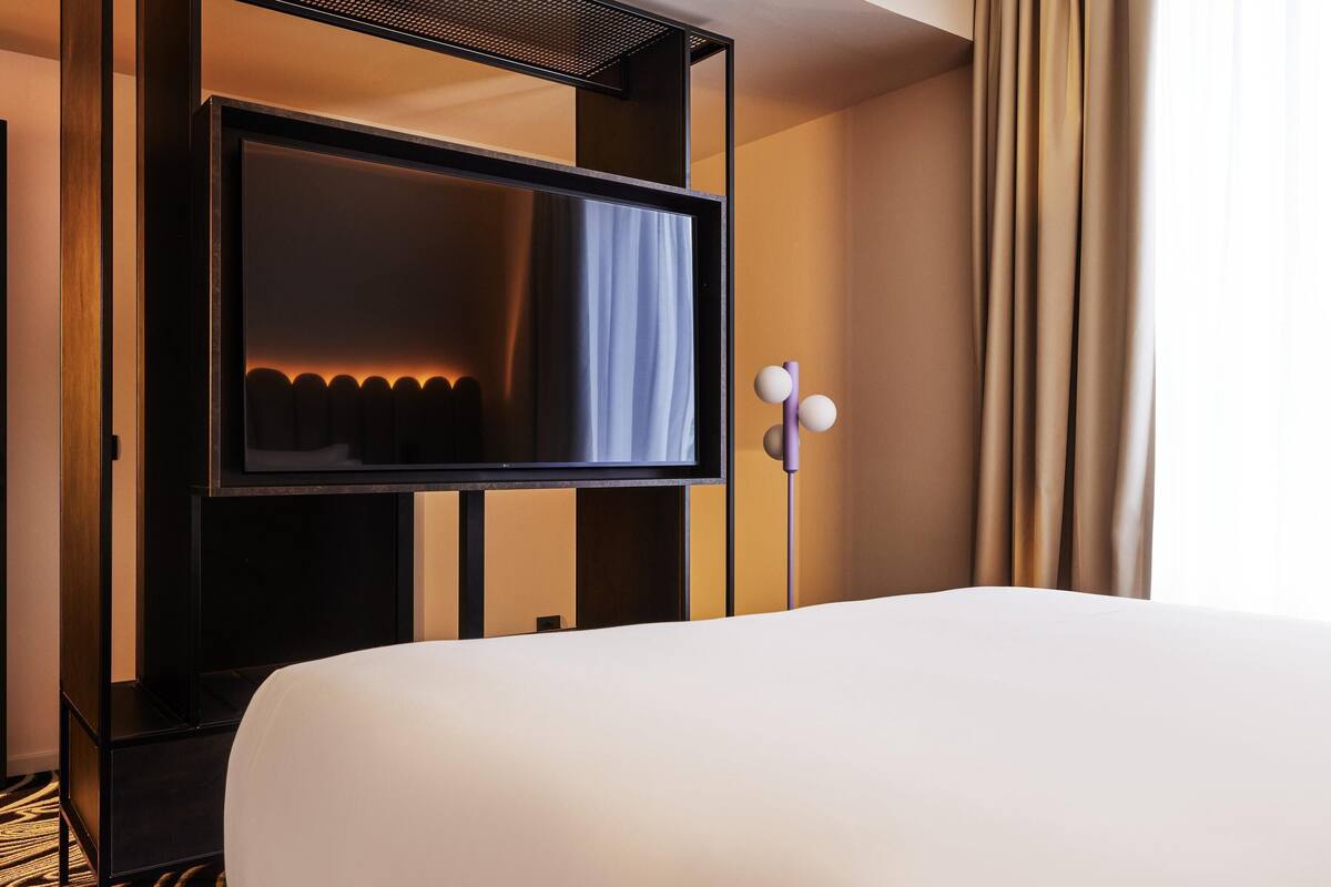 A hotel room with a bed, a television mounted on a black stand, and a curtain on the window.