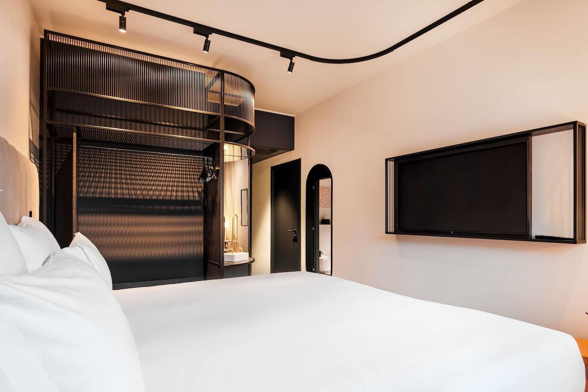 A modern hotel room with a large bed, a curved ceiling, and a built-in seating area with a view of the city.