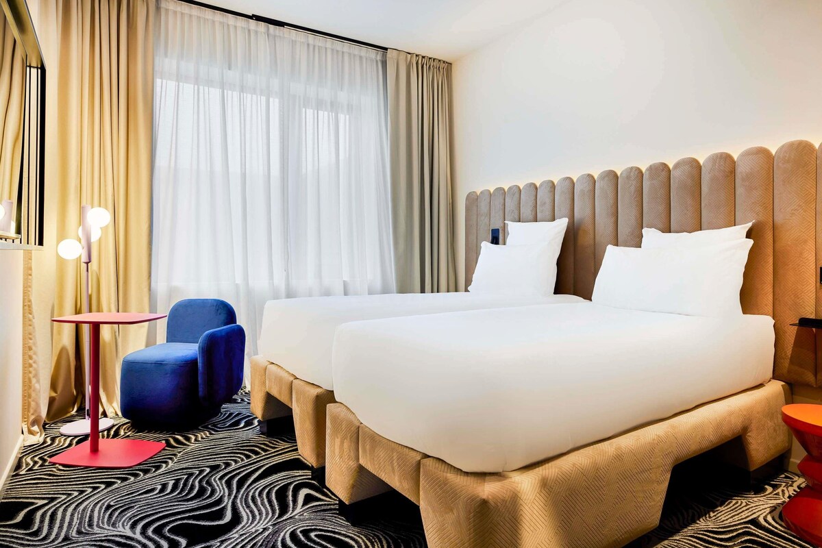 A hotel room with a bed, two bedside tables, a chair, and a patterned carpet.