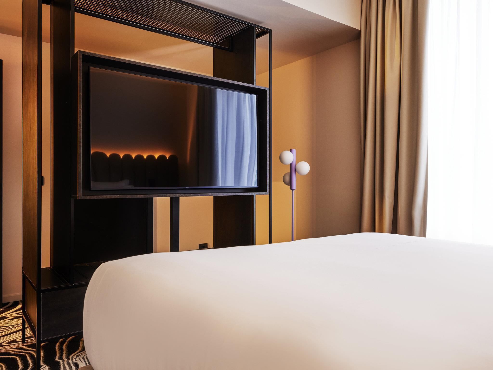 Une chambre d’hôtel avec un grand lit, un téléviseur à écran plat fixé sur un panneau en bois, une petite table et un rideau à la fenêtre.