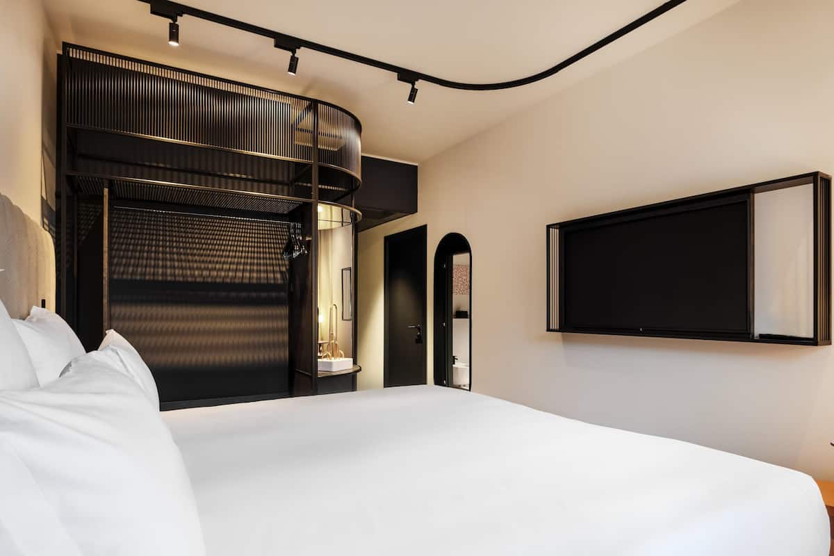 Une chambre dâhĂŽtel moderne dotĂ©e dâun grand lit, dâune tĂȘte de lit noire aux lignes courbes, dâun tĂ©lĂ©viseur fixĂ© au mur et dâun bureau intĂ©grĂ© avec une chaise.