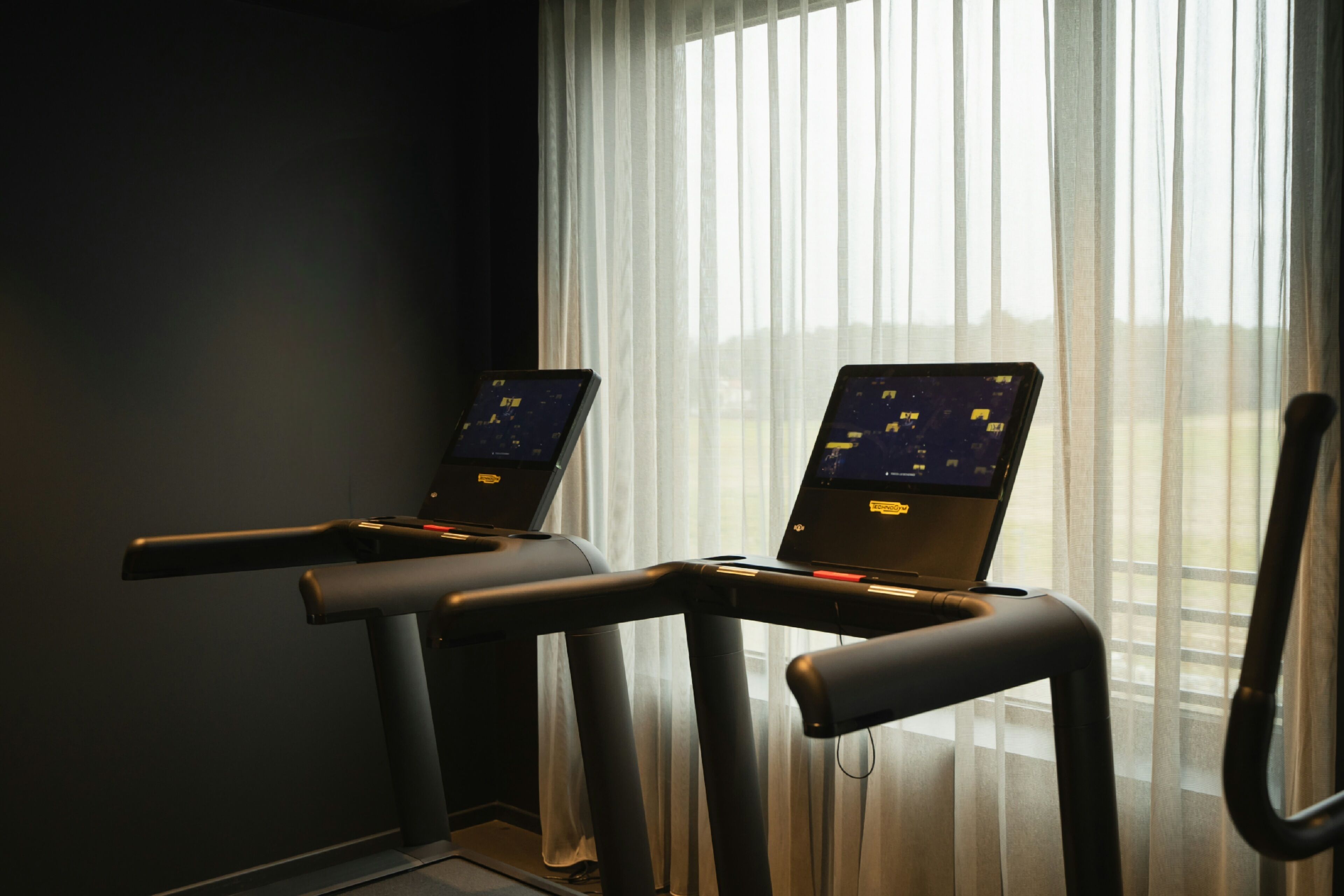 Two exercise machines with digital displays, positioned in front of a window with sheer curtains.