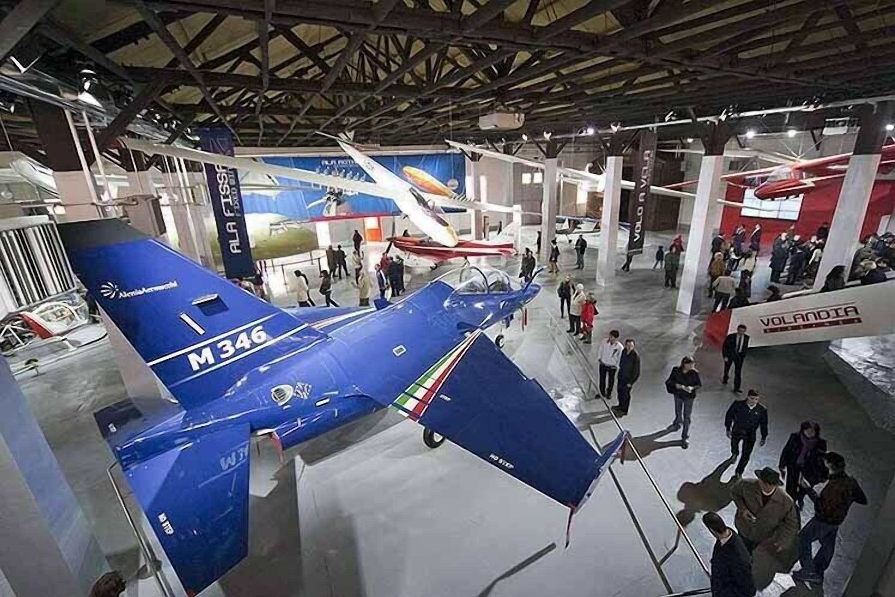 Una exhibición en interiores con aviones expuestos, incluyendo un avión azul con el número de registro M346.