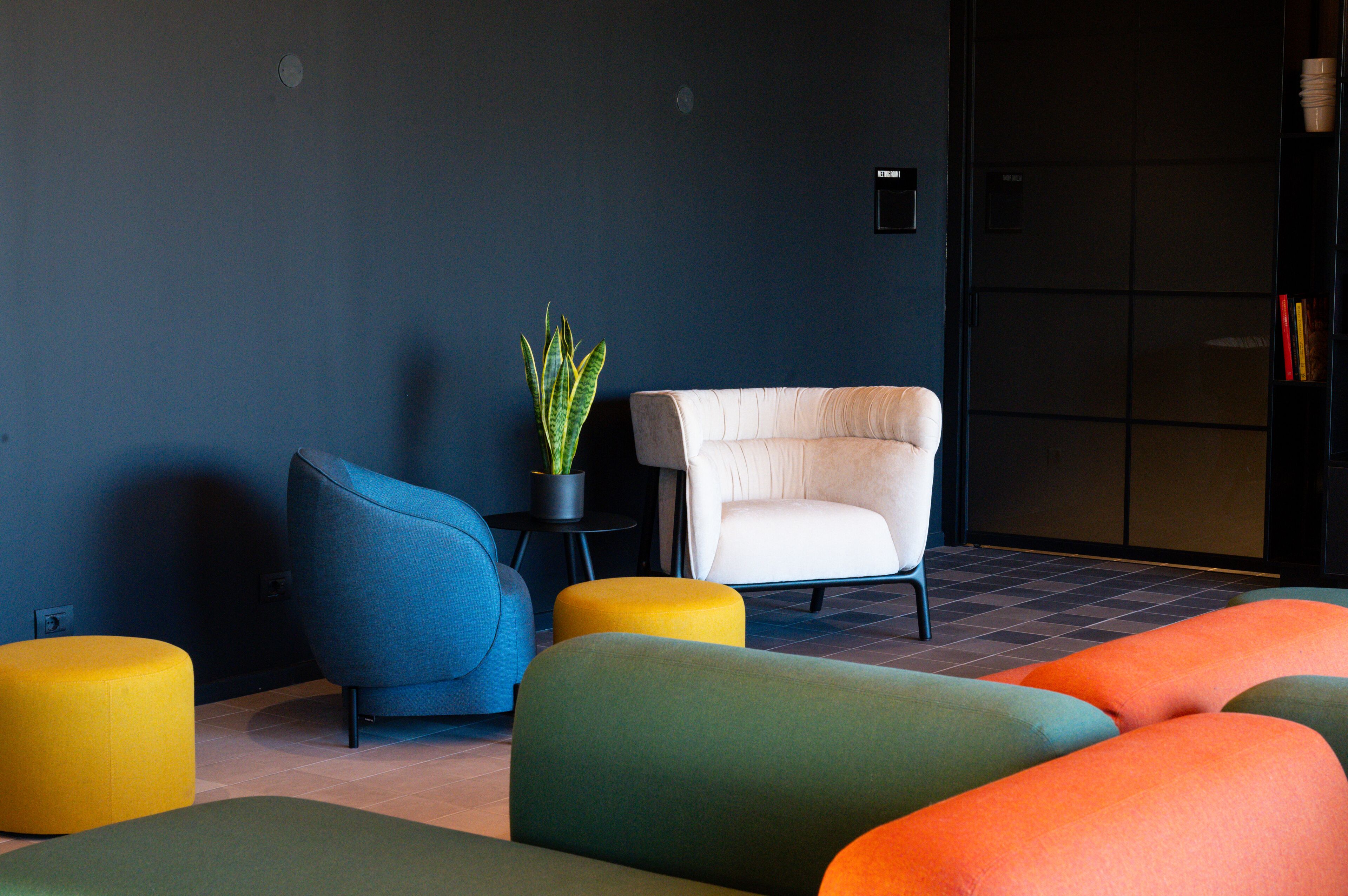 A modern living room with a dark blue wall, a white armchair, a blue sofa, and colorful ottomans.