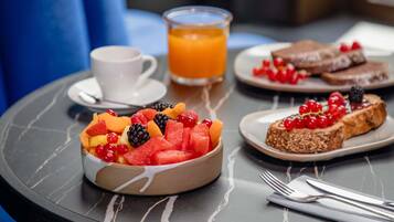 Une table mise pour le petit-déjeuner, avec un saladier de fruits, des toasts garnis de fruits rouges, une tasse de café et un verre de jus d’orange.
