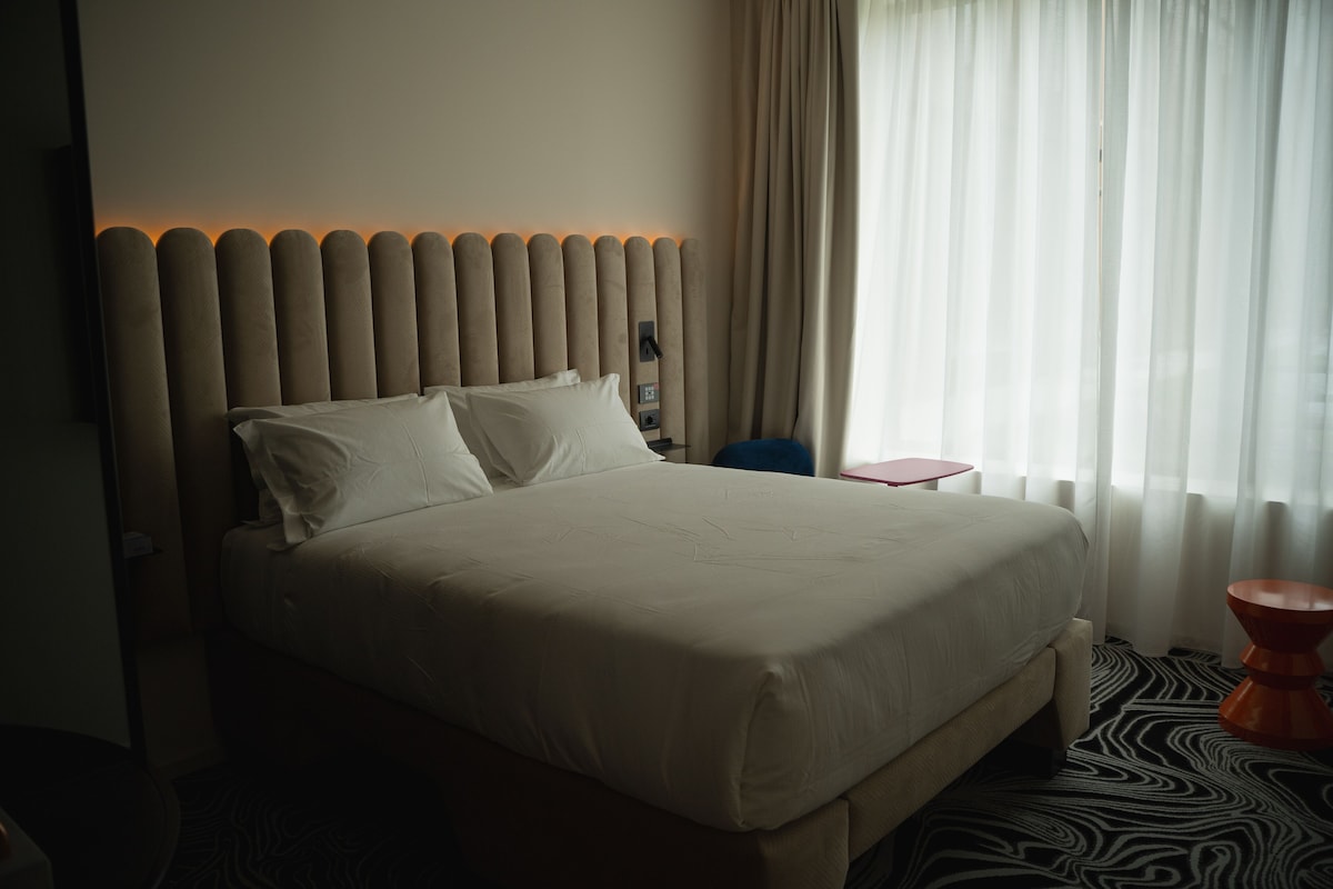 A hotel room with a large bed, a headboard with vertical panels, a small side table, and a window with sheer curtains.