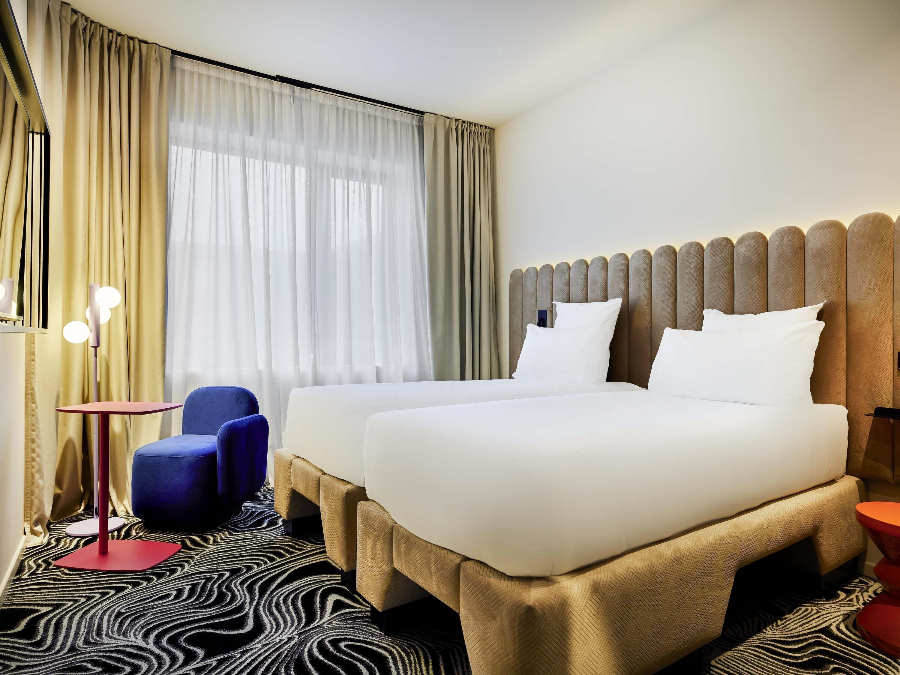 Une chambre d’hôtel avec deux lits, une chaise bleue, une table rouge et un tapis à motifs.