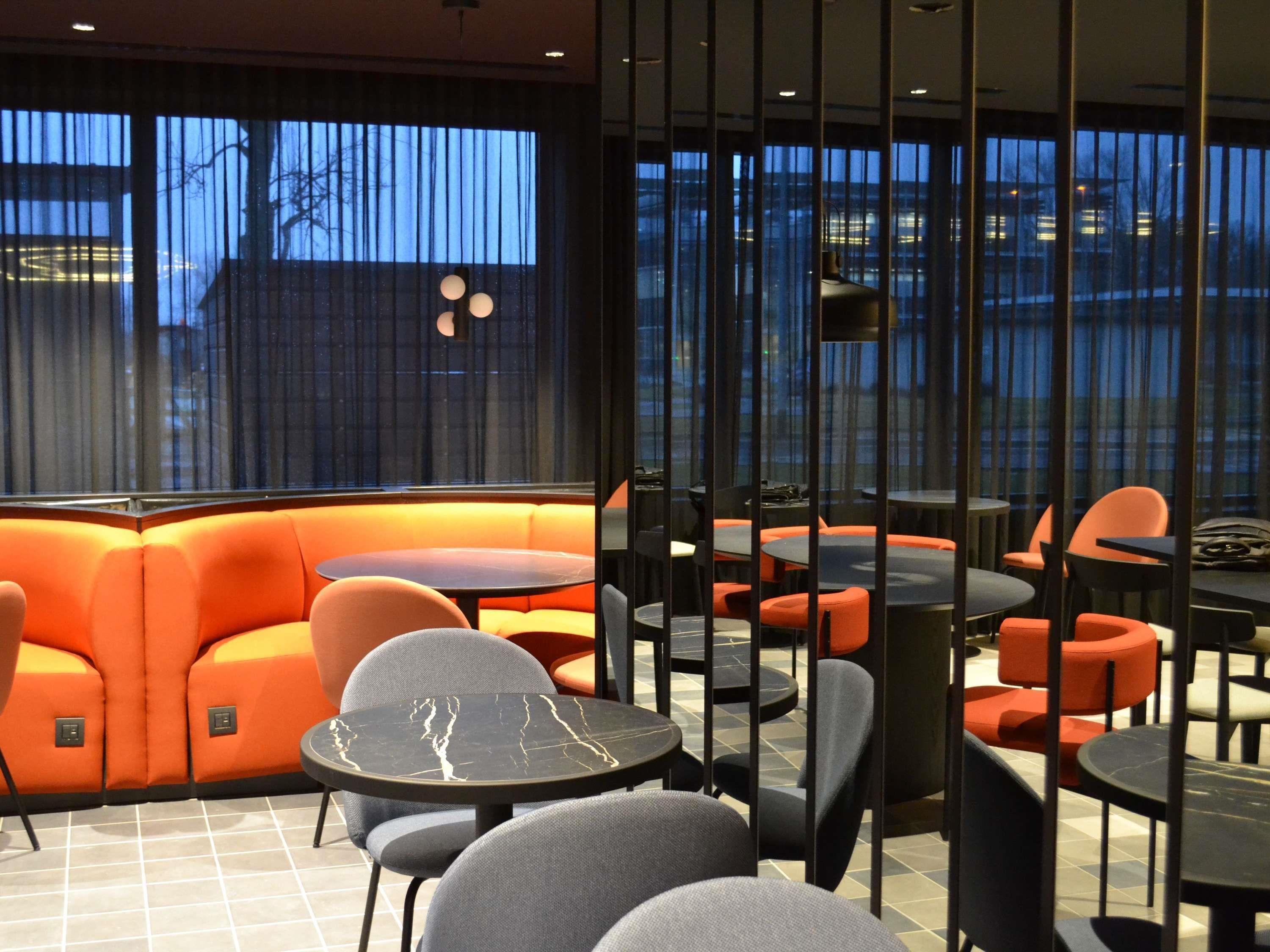 A modern interior with a seating area featuring orange sofas and round tables, large windows with a view of buildings, and a tiled floor.