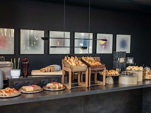 A buffet counter with various baked goods and pastries, a vase with yellow flowers, and framed abstract art on the wall. - Tribe Milano Malpensa (Vizzola Ticino)
