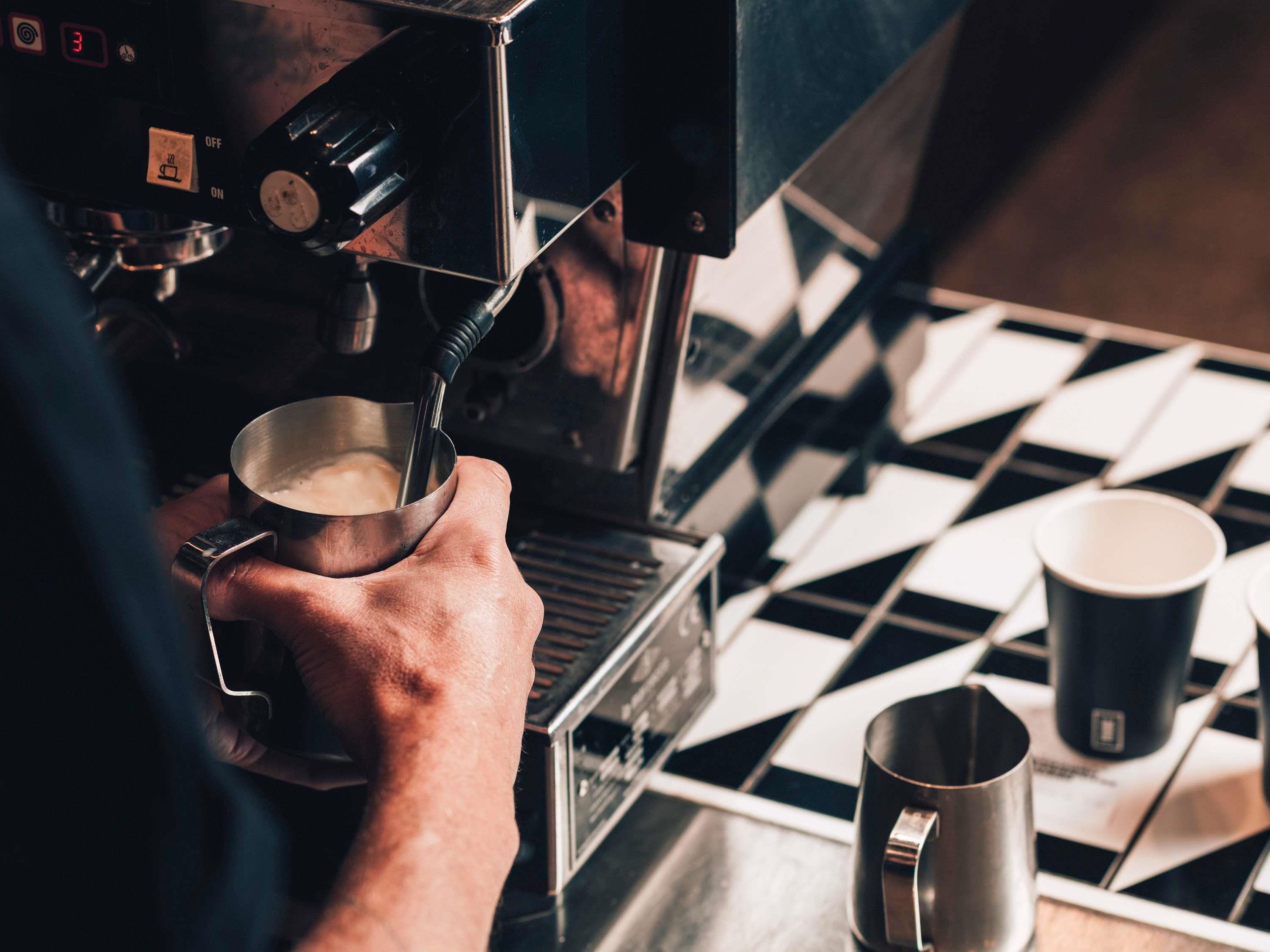 A person operating an espresso machine, holding a metal cup under the spout, with a checkered floor and two metal cups on the table.