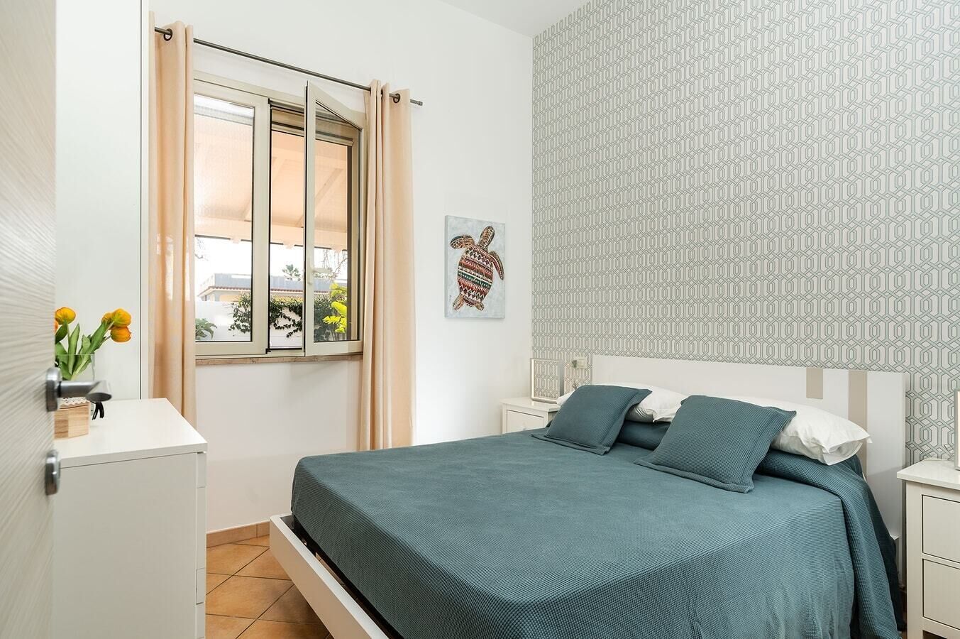 Un dormitorio con cama, mesita de noche, ventana con cortinas, pared con diseño geométrico y una planta pequeña en la mesita.