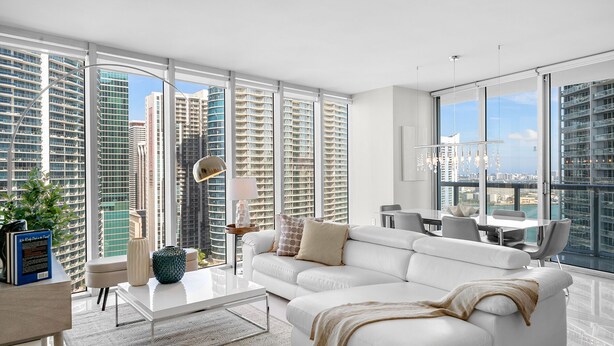 A modern living room with a white sofa, a coffee table, and a dining area with a view of tall buildings. - ICON W BRICKELL LUXURIOUS OCEAN VIEW 2BR/2BA #3002 (Miami)