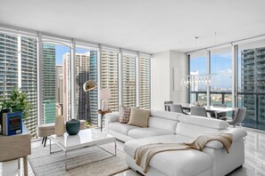 A modern living room with a white sofa, a coffee table, and a dining area with a view of tall buildings. - ICON W BRICKELL LUXURIOUS OCEAN VIEW 2BR/2BA #3002 (Miami)