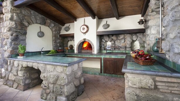 Una cocina rústica al aire libre con chimenea de piedra, vigas de madera y suelo de baldosas.