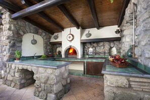 Una cocina rústica al aire libre con chimenea de piedra, vigas de madera y suelo de baldosas.