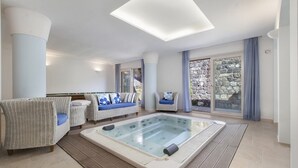 Habitación de spa con bañera de hidromasaje, dos sofás y un ventanal con vistas a una pared de piedra.