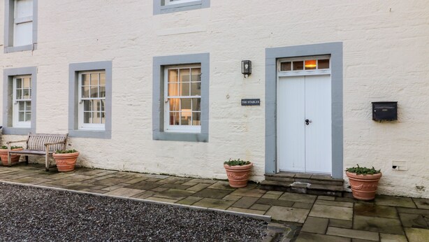 A white building with a white door, a bench, and potted plants. - The Stables (Dumfries)