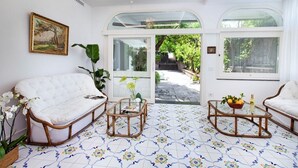A spacious living area with a white sofa, a coffee table, and a wicker chair. A large window offers a view of a garden.