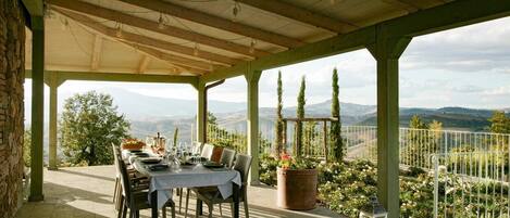 A covered outdoor dining area with a table set for four, overlooking a scenic landscape with mountains and trees.