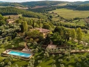 An aerial view of a property with a swimming pool, a large house surrounded by trees, and rolling hills in the background.